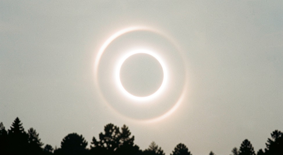 Un anneau lumineux pâle autour du soleil voilé dans un ciel de cirrostratus laiteux, silhouette de cimes en bas du cadre, lumière blanche.