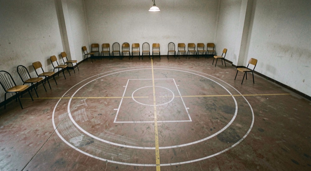 Un cercle tracé à la craie sur un sol de gymnase, des chaises repoussées contre les murs, lumière de plafonnier, vue de dessus, plan large.