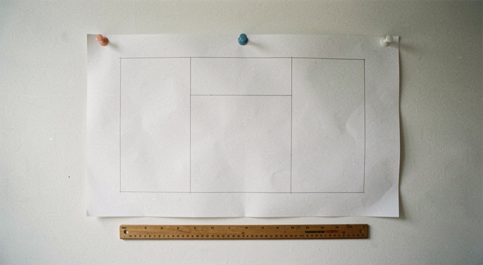 Une grande feuille blanche divisée en cinq rectangles tracés au crayon et punaisée sur un mur clair, règle posée dessous, lumière de studio, plan large.